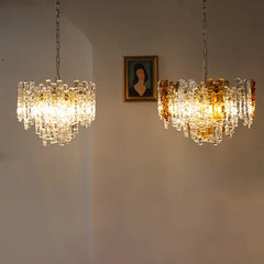Two glass tube chandeliers with amber and clear glass tubes hanging on chains against a neutral wall with a framed portrait above.