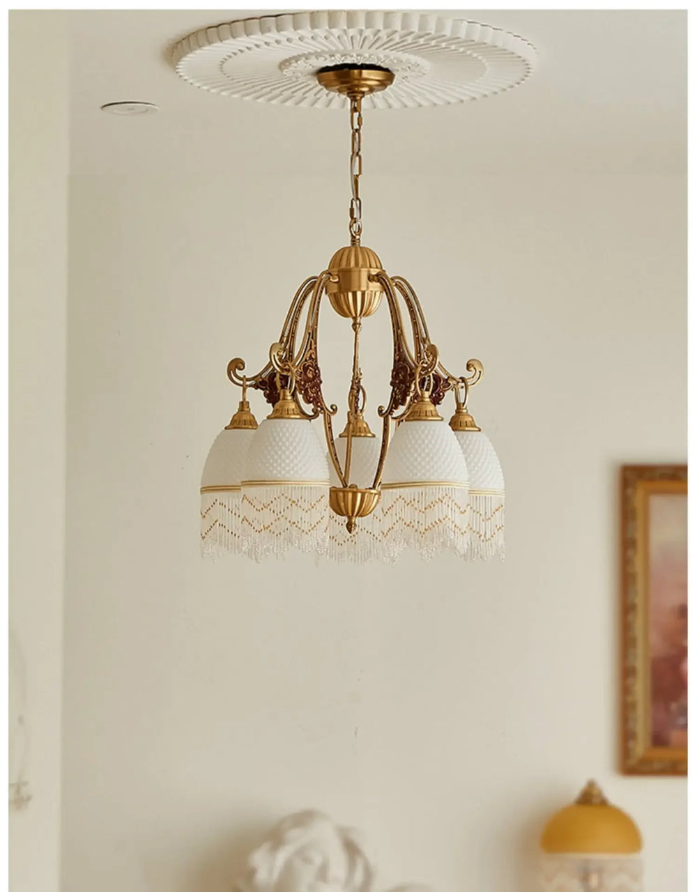 A polished brass chandelier with white glass shades and decorative fringe hangs from a white ceiling medallion, with a partial view of a sculpture in the foreground.