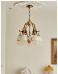 A polished brass chandelier with white glass shades and decorative fringe hangs from a white ceiling medallion, with a partial view of a sculpture in the foreground.