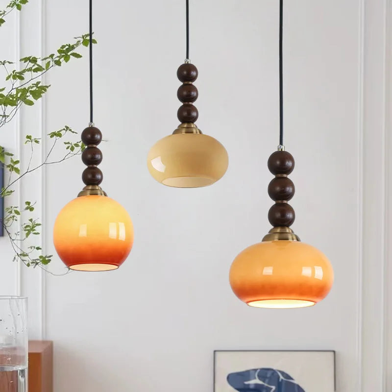 Three wood bead and persimmon-shaped globe pendant lights hanging against a white wall, with greenery on the left.