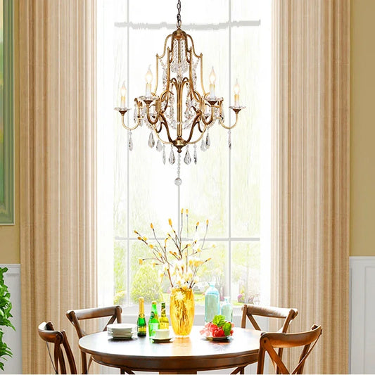 A gold candelabra crystal chandelier hanging above a round dining table in a room with beige curtains.
