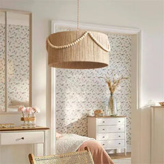 A bohemian-style rattan drum pendant light hangs in a softly lit room with patterned wallpaper and a dresser with flowers.