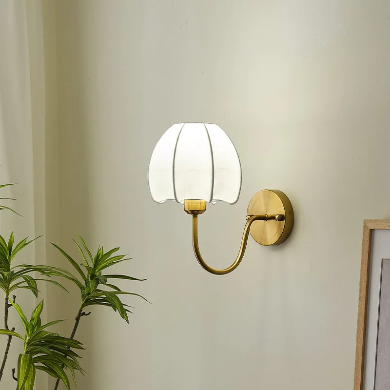A gold wall sconce with a white pleated fabric shade shaped like a flower, against a plain wall, with a framed picture partially visible to the right.