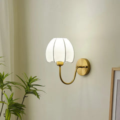 A gold wall sconce with a white pleated fabric shade shaped like a flower, against a plain wall, with a framed picture partially visible to the right.