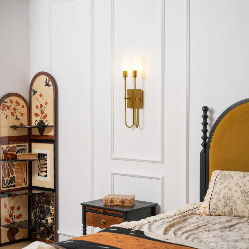 A brass-finished wall sconce with a double curved arm and frosted glass shades illuminates a modern bedroom beside a nightstand.