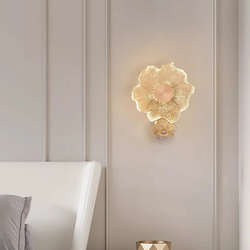 A gold flower glass wall sconce mounted on a light gray paneled wall next to a white headboard in a bedroom setting.