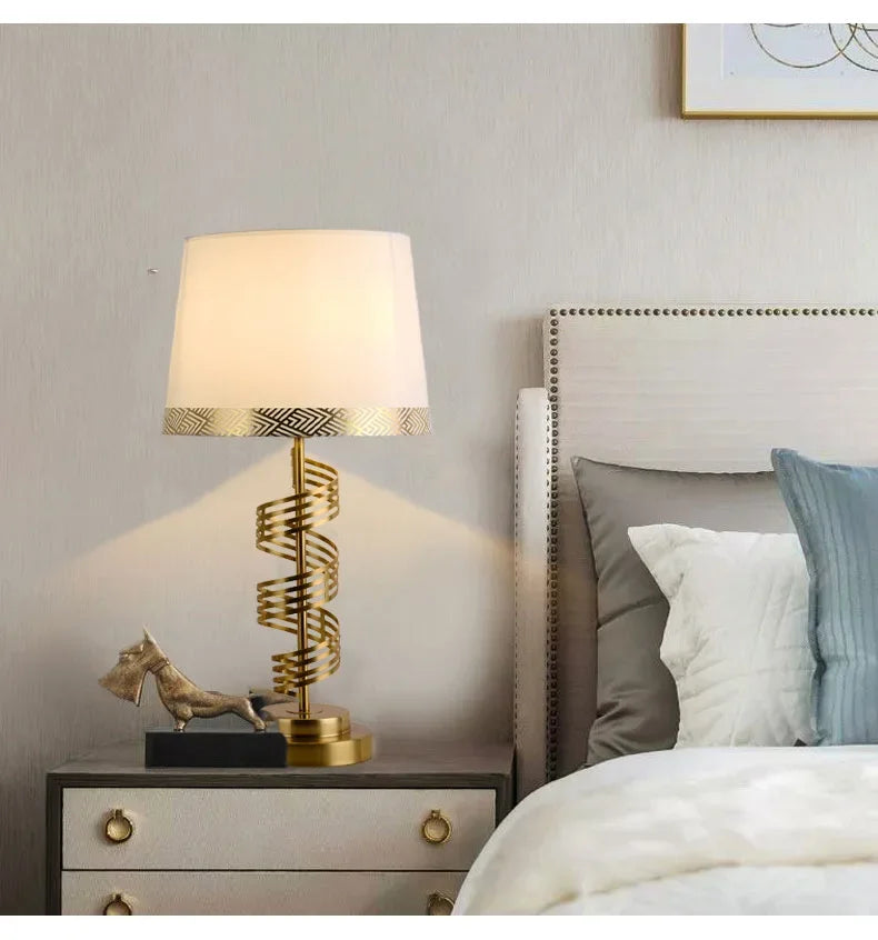 A gold spiral table lamp with a white conical fabric shade on a nightstand next to a bed.