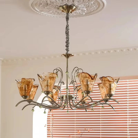 A wider shot of a bronze chandelier with amber glass shades and floral accents, illuminating a room with white blinds in the background.