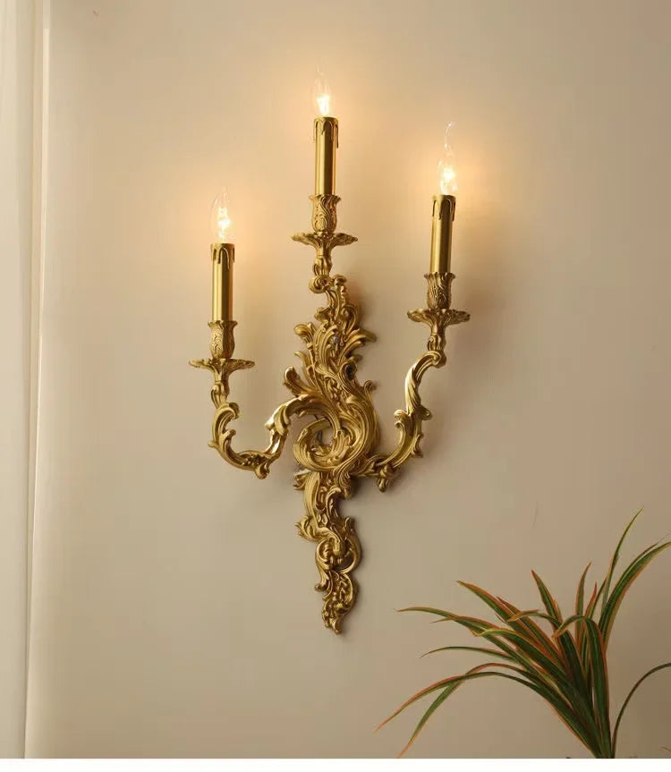 Side view of a gold candelabra wall sconce mounted on a wall, showing its decorative scrollwork and three illuminated candle lights.
