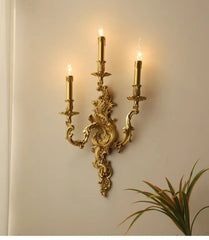 Side view of a gold candelabra wall sconce mounted on a wall, showing its decorative scrollwork and three illuminated candle lights.