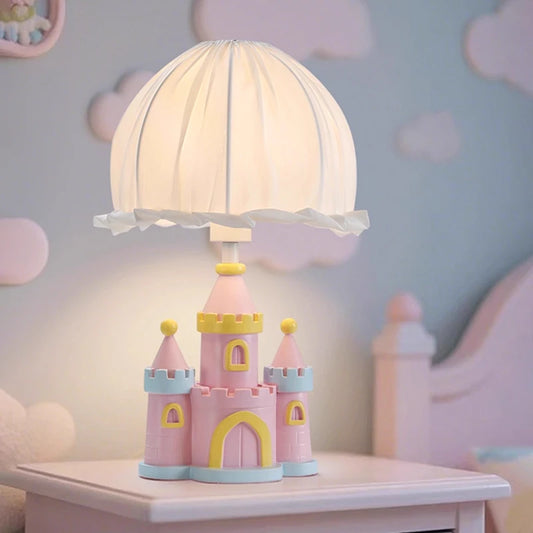 Pink castle-shaped table lamp with a pleated white fabric shade, set on a pink bedside table against a backdrop of pastel clouds.