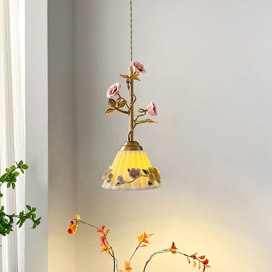 A single pendant light with a brass stem, pink rose buds, and a frosted glass shade with floral patterns, hanging against a white wall.