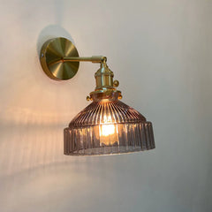 Pinkish-grey fluted glass shade wall sconce with a brass arm and mounting plate illuminated against a beige wall.
