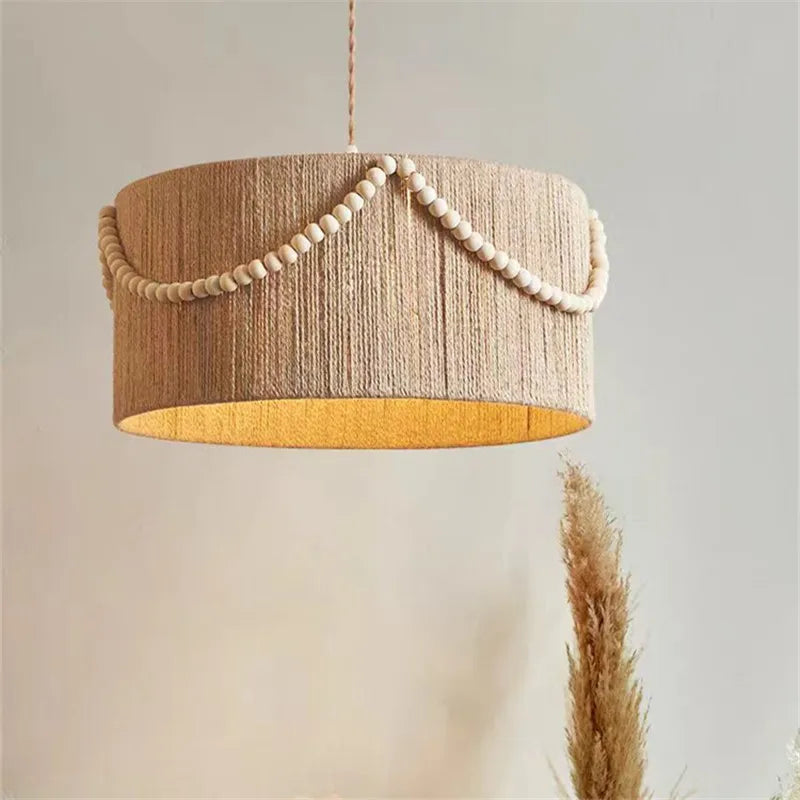 Close-up of a cylindrical pendant light fixture wrapped in natural-colored hemp rope, with a strand of small wooden beads draped along the top edge.