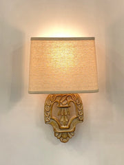 A vintage wood wall sconce with a carved base and fabric shade, illuminated against a white wall.