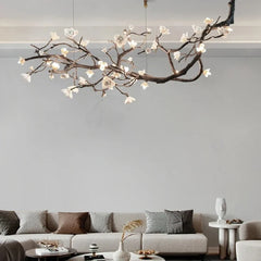 A long, artistic chandelier shaped like a branching tree with white floral accents and illuminated LED lights, hanging over a living room sofa.