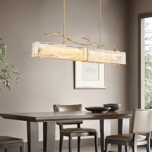 Rectangular glass pendant light with brushed gold branch accents hanging above a dining table.