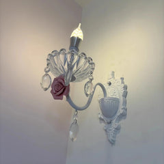 Close-up of the decorative elements of a white wall sconce, showing a pink ceramic rose and faceted crystal droplets attached to the light fixture.