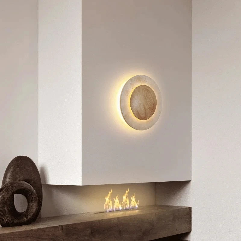 A round wall sconce with a natural stone finish and warm LED light, mounted on a white wall above a modern fireplace with a gas flame.