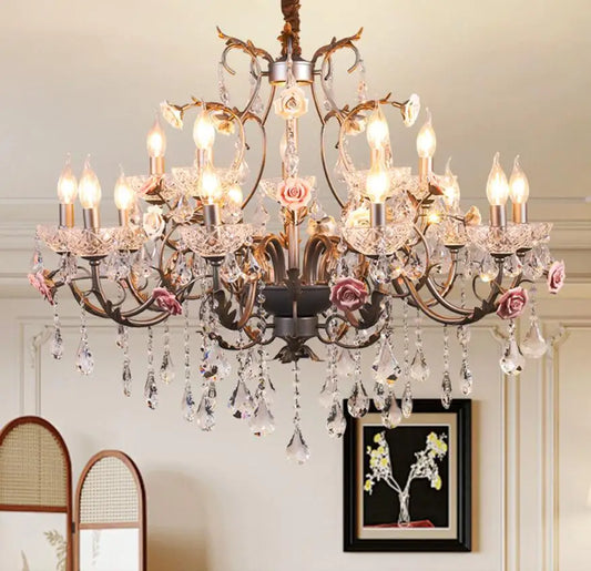 A close-up view of a gold-toned chandelier adorned with ceramic pink roses, cascading crystals, and multiple candle-style light sockets, emitting a warm glow.