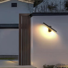 Black rotatable LED wall sconce mounted on an exterior wall, illuminating a pathway at night.