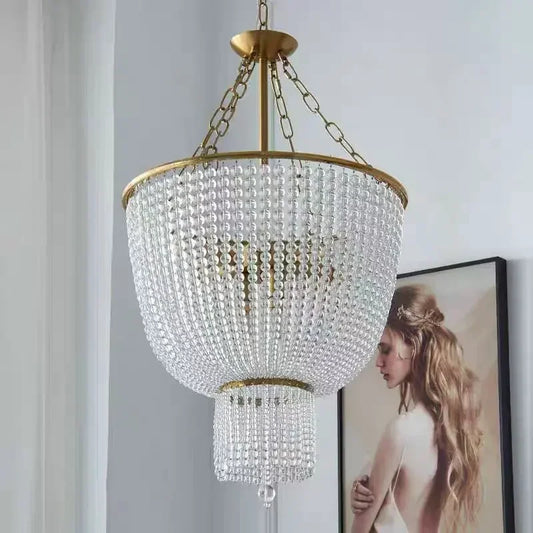 A gold-framed chandelier covered in clear crystals hangs in a bright room, near a framed portrait of a woman with long blonde hair.