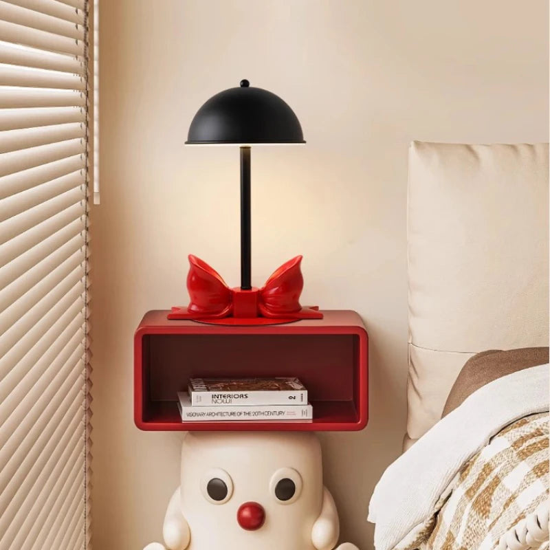 Red bow accent table lamp with a black dome shade mounted on a red shelf above a cartoonish figure.