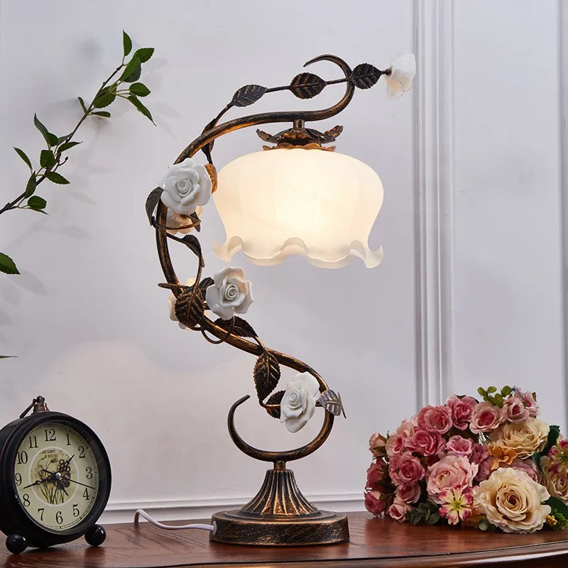 Decorative table lamp with floral design on a wooden surface next to a clock and flowers.