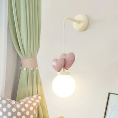 A wall sconce with a cream mounting bracket, a white cord, three pink heart-shaped accents, and a white globe light emitting bright white light.