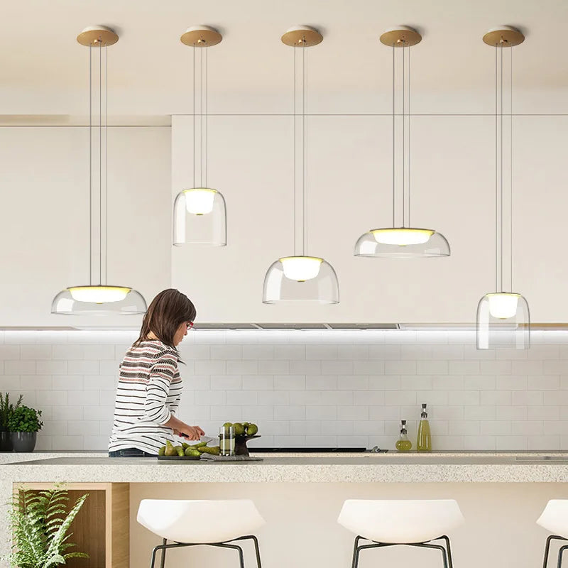 A woman in a kitchen preparing food, with five clear glass dome pendant lights with gold accents installed above the counter.