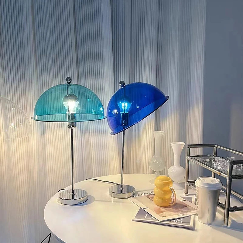 Two mushroom-shaped table lamps with blue and teal glass shades on a white table with white curtains in the background.
