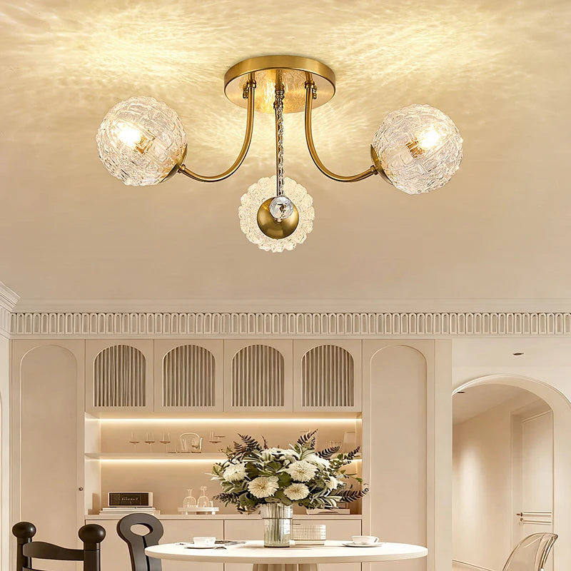 A polished gold semi-flush ceiling light with two ripple glass globes and a central crystal accent, illuminating a dining area with a table and chairs.