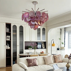 A modern chandelier with layered pink and purple glass discs hanging from a chrome frame, above a cream sofa in a well-lit room.