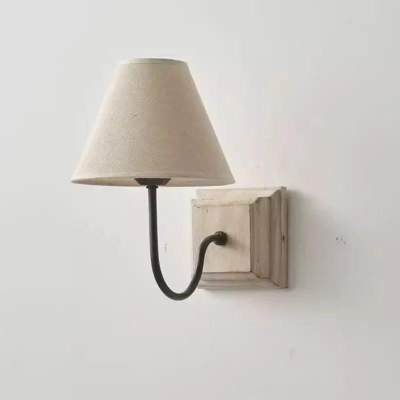 Side view of a wall sconce featuring a light beige fabric cone shade and a distressed white wood backplate, with a dark metal arm supporting the shade.