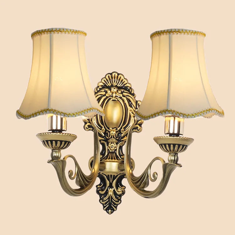 Frontal view of a two-light gold wall sconce with flared cream fabric shades, highlighting its elaborate metalwork.