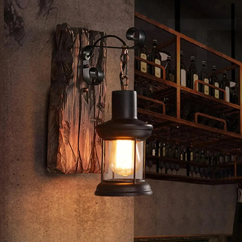 Black iron lantern wall sconce with a distressed wood backplate mounted on a wall next to a shelf unit filled with bottles.