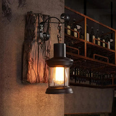 Black iron lantern wall sconce with a distressed wood backplate mounted on a wall next to a shelf unit filled with bottles.