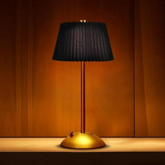 A black pleated fabric shade table lamp with a gold base and stem, illuminated against a warm wooden backdrop.