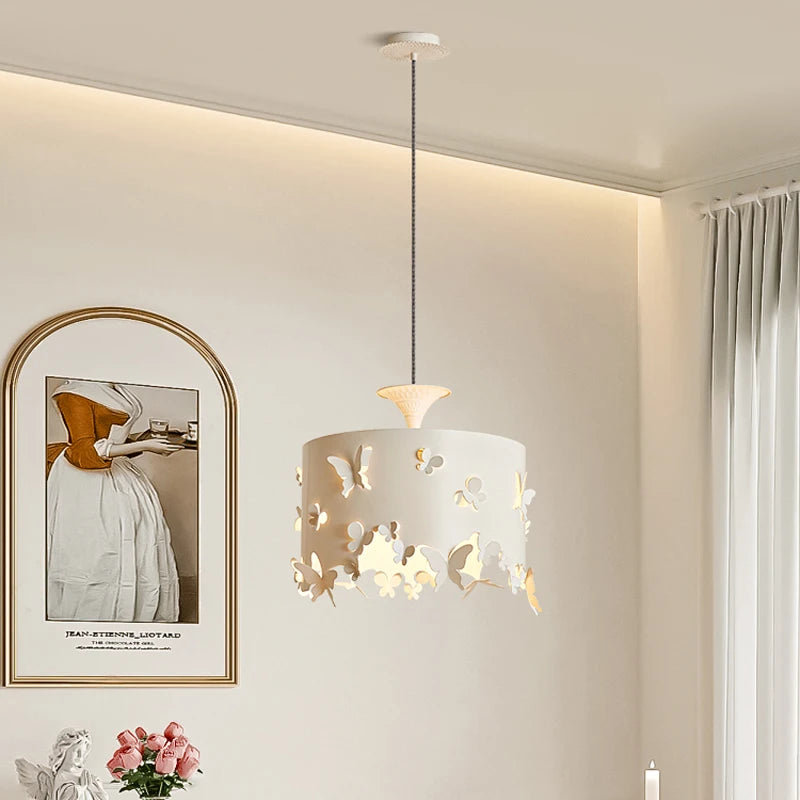 A cream-colored drum pendant light with intricate butterfly cutouts, hanging from a white ceiling. The light casts a warm glow through the cutouts.