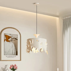 A cream-colored drum pendant light with intricate butterfly cutouts, hanging from a white ceiling. The light casts a warm glow through the cutouts.