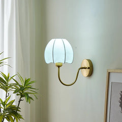 A gold wall sconce with a white pleated fabric shade shaped like a flower, mounted on a light-colored wall with a green plant in the foreground.