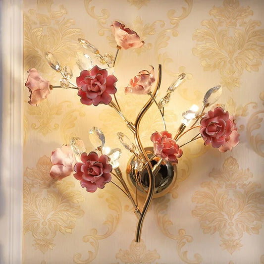 A gold wall sconce with multiple pink floral accents and clear crystal details, mounted on a textured wallpaper.