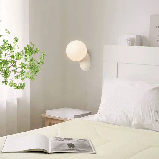 A minimalist wall sconce with a globe shade mounted above a bed in a bedroom setting.