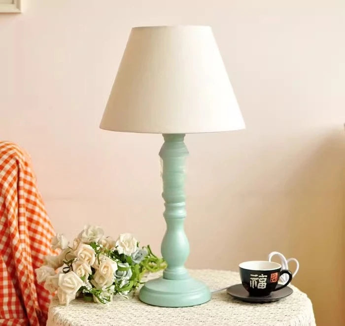 Light blue table lamp with white shade on a surface with flowers and a cup.