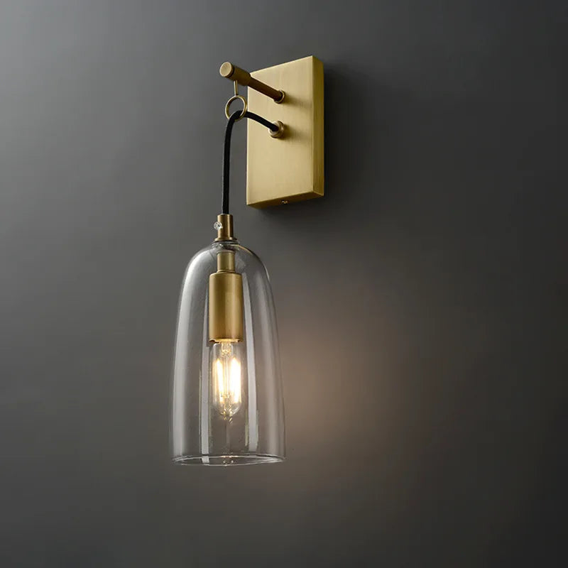 A modern gold and clear glass bell wall sconce mounted on a dark gray wall, with the light on.