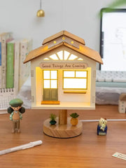 White wooden tree house shaped table lamp with a green door, windows, and a sign that reads 'Good Things Are Coming', illuminated by warm LED lights.