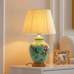 A turquoise ceramic table lamp with a white pleated fabric shade, decorated with painted flowers and butterflies, situated on a white nightstand.