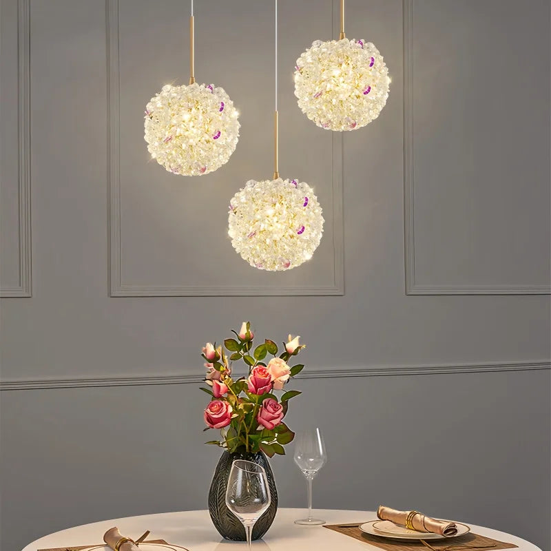 Three gold crystal orb pendant lights hanging over a dining table with a vase of roses.