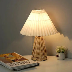 White pleated fabric shade table lamp with a woven rattan base on a nightstand next to a book.