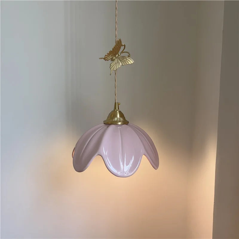 Illuminated pink ceramic petal pendant light with brass butterfly accent against a wall.